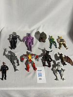 Overview photo showing eleven assorted 1980s action figures arranged on a white cloth background including a ninja figure, purple humanoid, green superhero, blond muscular man, and others.