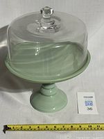 Front angle view of green jadeite cake stand with clear glass dome cover and measuring tape