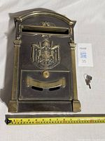 Front view of lockable metal mailbox showing decorative crest, mail slot and lockable front door with keyhole; key placed beside mailbox