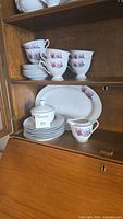 Full luncheon set with teacups stacked on saucers and side plates stacked below, sugar bowl with lid and creamer placed in front, and oval platter displayed at the back inside wooden cabinet.