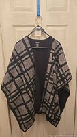 Black and gray plaid cape wrap hanging on a black hanger against a beige door. The cape has an open front and a relaxed fit.