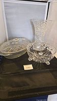 Photo showing three items: a crystal vase, a cut glass footed bowl, and an oval glass casserole dish with lid on a black surface against a white background.
