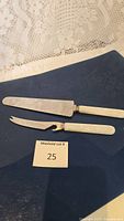 Two serving tools laid on a dark surface with a lot number card. Tools have stainless steel blades and smooth white faux pearl handles.