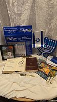 Jewish cultural items including blue LED electric menorah in box, paper menorah decoration, Israeli flags, two high holiday prayer books, and multiple mezuzah cases on a white cloth.