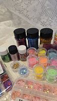 Photo showing multiple small sealed plastic bottles of glitter in purple, blue, gold and small plastic containers of neon colored powders and embellishments on a lace background.
