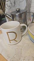 Photo of white ceramic mug with gold letter 'R' alongside stainless steel teapot and some plates