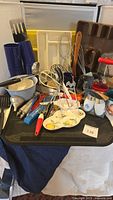 Full overview of miscellaneous kitchen utensils and baking items showing a variety of spatulas, whisks, trays, cups, bowls, and molds stacked on a black plastic chair.