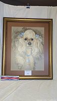 Framed pastel drawing of white poodle with blue bows and pendant, hung for display to show entire frame and artwork