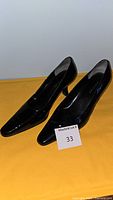 Pair of black leather women's pumps shown side by side from a front-side angled view on a yellow surface with lot number card.