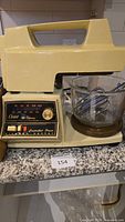 Vintage Oster Kitchen Center stand mixer in cream-yellow with glass mixing bowl and control panel with dials and switches shown on granite counter.