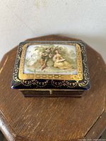 Top view of the trinket box showing the hinged lid with detailed painted scene of a courting couple and gold decorative border.