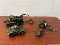Four toy military vehicles on wood surface including truck, armored car, artillery, and boxy artillery piece.