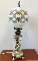 Full view of the lamp showing the white painted glass balloon shade with gold and black patterns, metal cherub base and hanging glass prisms.