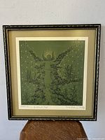 Framed colored lithograph depicting a central glowing figure with stars and intricate village houses on a green background.