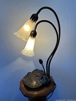 Lamp illuminated showing two lily-shaped art glass shades lit, attached to curved metal stems on a lily pad-style metal base.