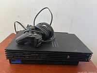 Front angle view of Sony PlayStation 2 console with original wired controller coiled on top, showing power and eject buttons and the blue USB port on the front left.