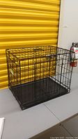 Black metal wire folding dog crate with plastic bottom tray, displayed on gray table against yellow background.