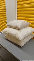 Stack of three cushions: two large fuzzy off-white Euro cushions and one beige geometric cushion on top.