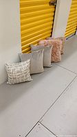 Five toss cushions, three gray solid, one pink textured, one gray-white pattern with metallic accents, lined up on floor.