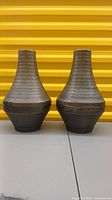 Pair of metal vases with textured geometric pattern and tapered necks on a grey surface against a yellow background.