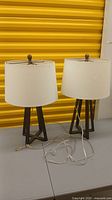 Two dark bronze table lamps with white shades side by side on gray surface in front of yellow wall.