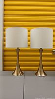Front view of both gold table lamps with white drum shades in front of yellow background.