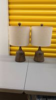 Pair of wooden lamps side by side on table against yellow background, showing lamp bases and beige fabric shades.