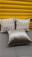 Set of three silver-themed toss cushions: two with white linen-look fabric and silver geometric embroidery, one in silver/gray textured velvet, displayed against a gray surface and yellow corrugated background.