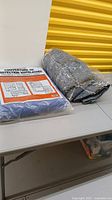 Photo showing one gray padded moving blanket rolled up and two blue felt moving blankets, one in plastic packaging.