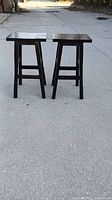 Photo shows two black wooden stools side by side on outdoor pavement, displaying front and side views.