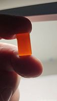Close-up photo of a rectangular cushion cabochon cut carnelian gemstone held between fingers against backlight, showing its vibrant mandarin orange color and translucency.