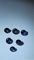 Photo of six round cut blue sapphires arranged on a flat surface, showing their size and color under lighting.