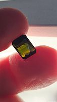 Close-up photo of a transparent bright green octagon cut tourmaline gemstone held between fingers against backlight showing color and facets clearly.