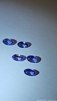 Top-down view of five oval cut purple-blue tanzanite gemstones arranged on a white surface showing their color and faceted cuts.
