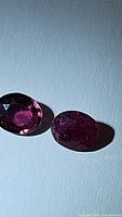Two oval-cut natural pink purple tourmaline gemstones, one with visible inclusions and the other cleaner, placed on a textured surface under natural light.