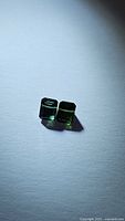 Pair of small fancy cut green tourmaline gemstones shown on white surface with light shadows, showing color and cut detail.