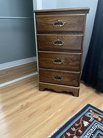 Tall narrow oak dresser with four drawers and brass handles, sitting on wooden floor next to black curtain and wall.