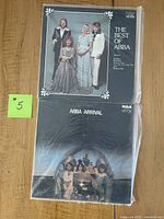 Two Abba vinyl albums displayed flat on a wooden surface. The top album is 'The Best of Abba' featuring a color photo of the band in classic 70s attire on a black background. Below is 'Abba Arrival' with the band inside a helicopter. Both albums are in sleeve covers and appear to be in used but good condition.