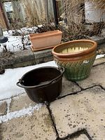 Three assorted outdoor pots displayed on a stone patio with some snow and dried plants in the background.