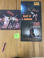 Photo of three vinyl album covers on a wooden surface: Ozzy Osbourne's Diary of a Madman and Blizzard of Ozz, and Judas Priest's Unleashed in the East. A pen and a small green note marked '#20' are also visible.