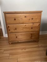 Front view of a natural pine wood dresser with three drawers and round wooden knobs set on ball feet. Measures approximately 31.5" wide, 17.5" deep, and 31" high.