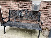 Black rod iron outdoor bench showing detailed decorative backrest with birds and vines, placed against a brick wall outdoors.