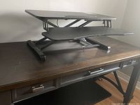 Black adjustable standing desk converter on top of wooden desk, showing side view with scissor lift mechanism