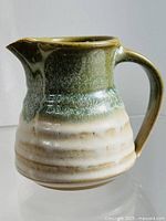 Front angle showing the curving spout, handle, and color gradient from green to cream on the pottery creamer.