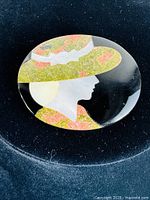 Full view of Victorian Pietra Dura brooch with profile of woman wearing a decorated hat with bow, made with inlaid green and pink stones and white shell on black background.