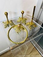 Four vintage cast brass bathroom fixtures including towel ring, two wall hooks, and toilet paper holder on clear acrylic stand