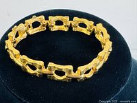 Full view of the gold tone bracelet displayed on a black cushion showing textured rectangular links and clasp.
