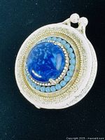 Close-up front view of circular ceramic pin box with large blue center stone surrounded by turquoise stones and textured off-white ceramic body with gold-toned trim and clasp.