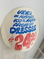 Close-up photo of front of circular sign showing hand-painted text advertising ladies or misses 100% rayon fashion dresses priced at $24.99.