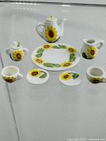 Full set displayed on clear surface showing all items separately: teapot, creamer, sugar bowl, two cups, two saucers, and plate with sunflower design.